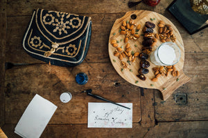 Camel milk on a wooden serving board with dates and nuts. There is a card on the table that says "Ramadan Kareem" written in calligraphy. There is a an ornate blue hand bag with a golden pattern on the table as well.