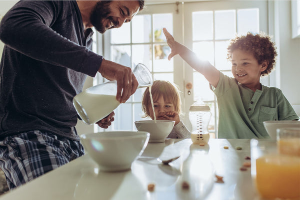 Man pouring milk into a bowl with a child reaching out to touch it, in a kitchen setting.