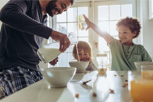 Man pouring milk into a bowl with a child reaching out to touch it, in a kitchen setting.