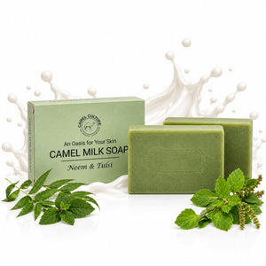 Two rectangular bars of Camel Milk Soap in a natural green shade are displayed in front of their light green packaging box. Fresh green neem and tulsi leaves are arranged around the base of the soap bars, with a stylized white splash graphic in the background.